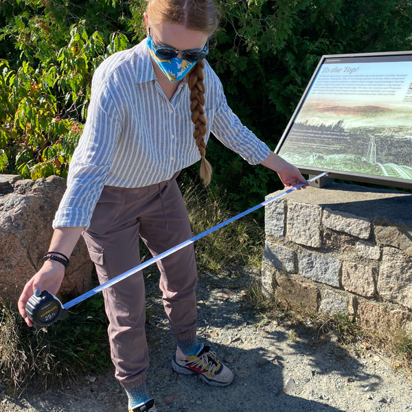 A woman holding a measuring tape near a park wayside exhibit
