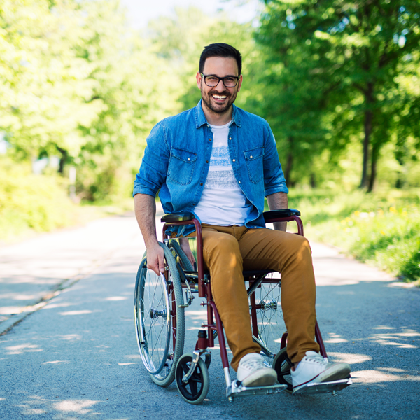A smiling man in a wheelchair on an outdoor paved path surrounded by trees
