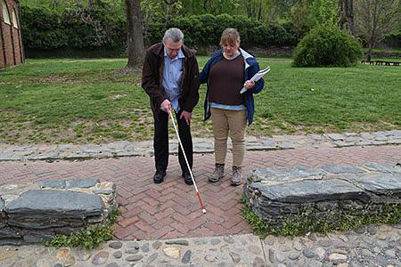 A blind man with a cane, assisted by a woman, on a park paved path